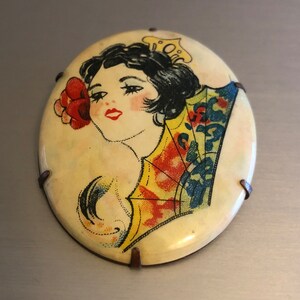 Pu&ograve; includere: Broche vintage di forma ovale con un'immagine colorata di una donna con un ventaglio e un fiore. La broche &egrave; in metallo con sfondo bianco e ha quattro punte metalliche sul retro.