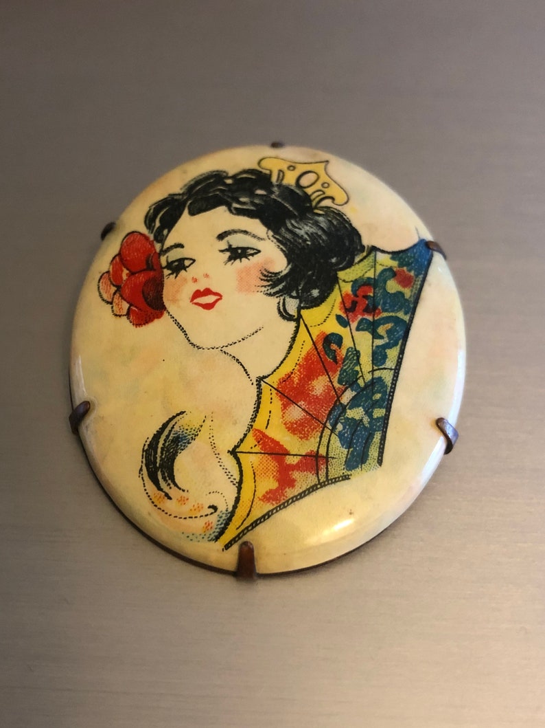 Pu&ograve; includere: Broche vintage di forma ovale con un'immagine colorata di una donna con un ventaglio e un fiore. La broche &egrave; in metallo con sfondo bianco e ha quattro punte metalliche sul retro.