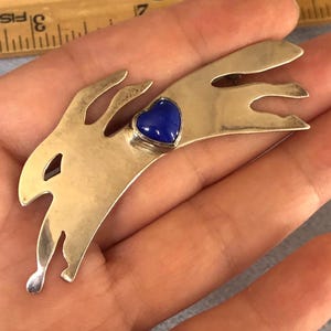 May include: A silver-coloured metal brooch shaped like a running rabbit. It has a heart-shaped, deep blue gemstone in the middle. The design is stylized and has a polished finish.