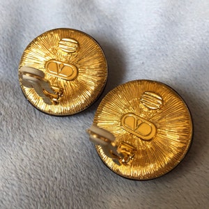 May include: A pair of gold-tone clip-on earrings with a black circular stone and a raised, textured design. The back of the earrings feature the Valentino logo.