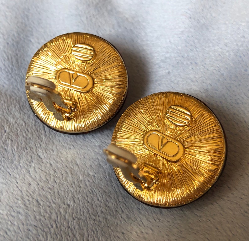 May include: A pair of gold-tone clip-on earrings with a black circular stone and a raised, textured design. The back of the earrings feature the Valentino logo.