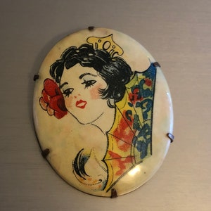 Pu&ograve; includere: Uno spillo vintage di forma ovale con un'immagine colorata di una donna con un fiore nei capelli e un ventaglio dietro di lei. Lo spillo ha uno sfondo bianco ed &egrave; circondato da una cornice metallica con piccoli accenti metallici.