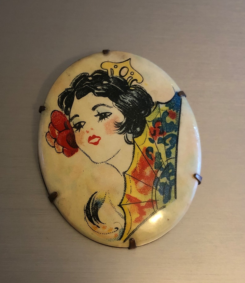 Pu&ograve; includere: Uno spillo vintage di forma ovale con un'immagine colorata di una donna con un fiore nei capelli e un ventaglio dietro di lei. Lo spillo ha uno sfondo bianco ed &egrave; circondato da una cornice metallica con piccoli accenti metallici.