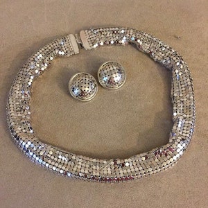 Puede incluir: Un collar y pendientes a juego de color plateado. El collar está hecho de una cadena de pequeños eslabones cuadrados. Los pendientes son redondos y tienen un diseño similar al del collar.