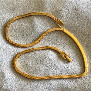 May include: A gold-colored necklace with a snake chain design. The necklace has a rectangular clasp and a curved bar. The chain is made of small, interlocking links. The necklace is displayed on a light gray surface.