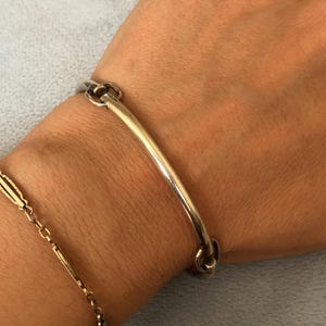 May include: A silver-colored bracelet with a curved bar design and two circular clasps. A gold-colored chain bracelet is also visible. The jewelry is displayed on a light blue surface.