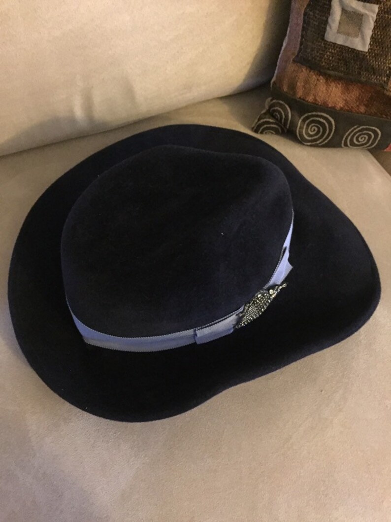 May include: A navy blue felt fedora hat with a light blue ribbon and a silver embellishment.