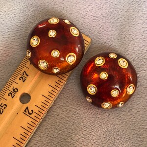 May include: A pair of brown and gold tone earrings with clear rhinestones. The earrings are round and have a textured surface.