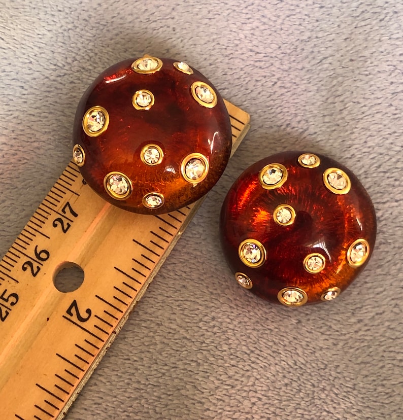 May include: A pair of brown and gold tone earrings with clear rhinestones. The earrings are round and have a textured surface.