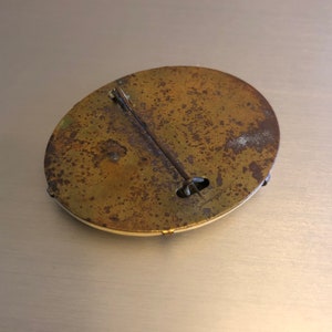 Pu&ograve; includere: Il retro di una spilla rotonda in ottone con uno spillo e una chiusura. La spilla &egrave; opaca e ha una finitura usurata.