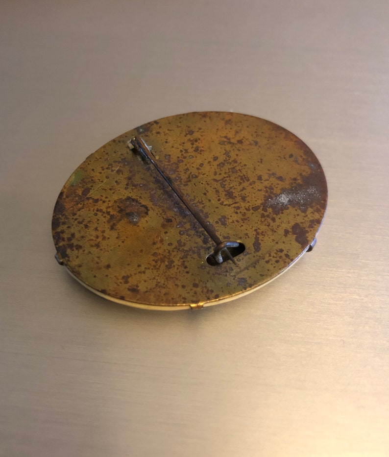 Pu&ograve; includere: Il retro di una spilla rotonda in ottone con uno spillo e una chiusura. La spilla &egrave; opaca e ha una finitura usurata.
