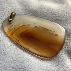 May include: A polished, teardrop-shaped pendant with a translucent, layered design. The pendant features swirling patterns of brown and white, with a brass-colored bail for attaching to a chain. The stone is approximately 2 inches long.