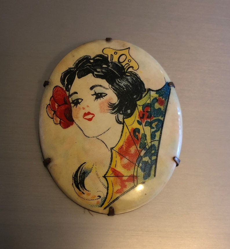 Pu&ograve; includere: Specchio compatto vintage di forma ovale con un'immagine colorata di una donna con un fiore nei capelli e un ventaglio. La donna indossa un vestito a motivi gialli e rossi. Lo specchio ha uno sfondo bianco ed &egrave; decorato con accenti metallici.