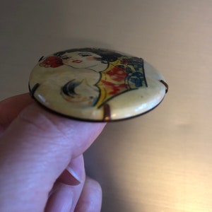 Pu&ograve; includere: Uno spillo rotondo di colore crema con un'immagine colorata di una donna con un fiore rosso nei capelli. Lo spillo ha un retro in metallo con due anelli di filo.