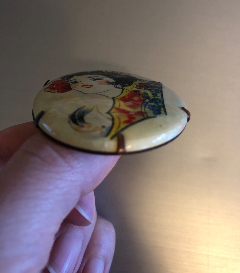 Pu&ograve; includere: Uno spillo rotondo di colore crema con un'immagine colorata di una donna con un fiore rosso nei capelli. Lo spillo ha un retro in metallo con due anelli di filo.