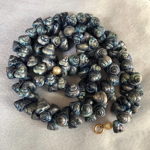May include: A necklace made from small, spiralled seashells in blue and silver hues. The shells are strung together, with a pearl bead and a gold-coloured clasp. A distinctive piece of jewellery.