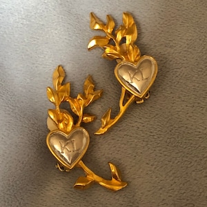 Puede incluir: Dos broches de corazón de tono dorado con un centro de tono plateado. Cada corazón tiene las iniciales "KL" grabadas en la parte delantera. Los broches están unidos a un tallo dorado con hojas.