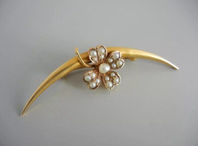 May include: A gold brooch with a crescent-shaped bar and a four-leaf clover design set with pearls.