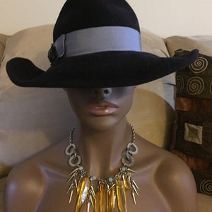 May include: A black wide-brimmed hat with a gray ribbon and a decorative embellishment. The hat is worn by a mannequin with a gold and amber necklace.