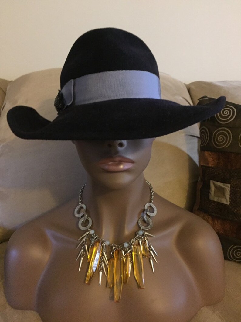 May include: A black wide-brimmed hat with a gray ribbon and a decorative embellishment. The hat is worn by a mannequin with a gold and amber necklace.