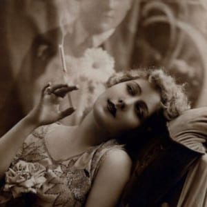 May include: A woman in a beaded dress with a floral embellishment is reclining on a couch, holding a cigarette in her hand. The image is in black and white.