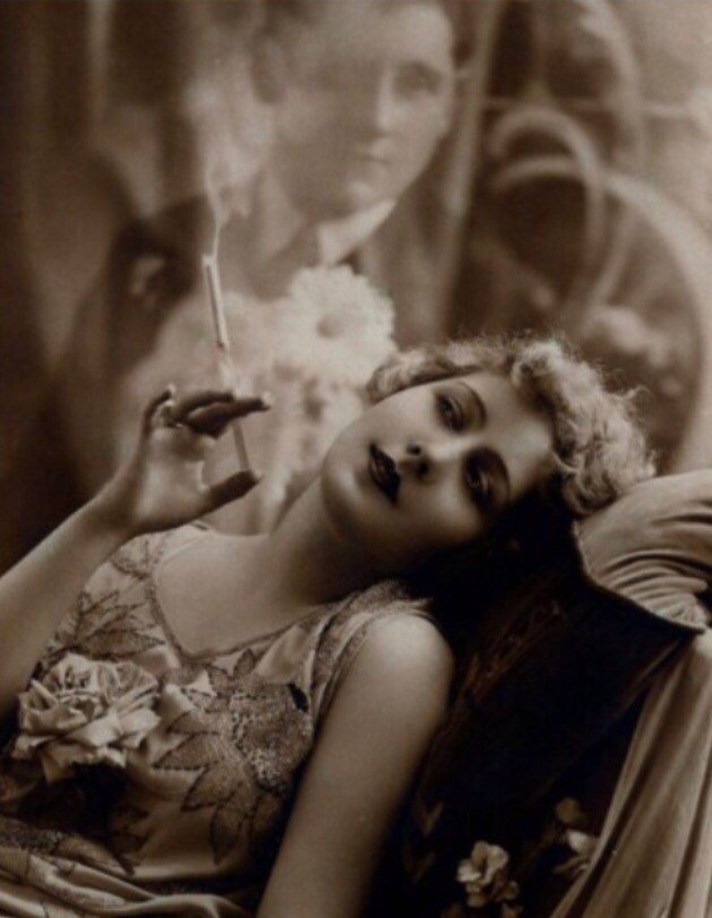 May include: A woman in a beaded dress with a floral embellishment is reclining on a couch, holding a cigarette in her hand. The image is in black and white.