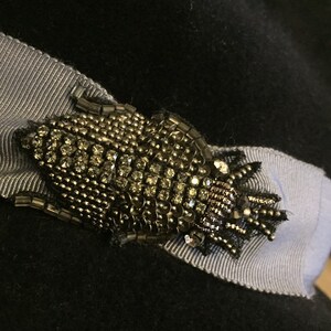 May include: A black hat with a gray ribbon and a gold and black beaded brooch. The brooch is shaped like a bug with a crown on its head.