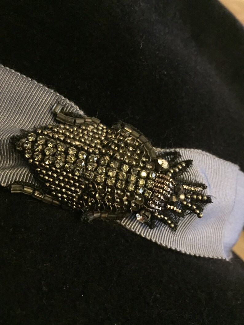 May include: A black hat with a gray ribbon and a gold and black beaded brooch. The brooch is shaped like a bug with a crown on its head.