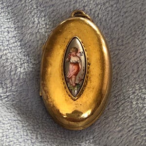 May include: A gold oval locket with a painted image of a cherub inside. The cherub is wearing a pink dress and has a white halo. The locket has a detailed design around the edge.