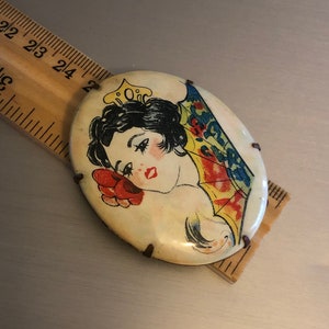 Pu&ograve; includere: Una spilla vintage di forma ovale con un'immagine colorata di una donna con una corona e un ventaglio. La spilla &egrave; in metallo e ha uno sfondo bianco.