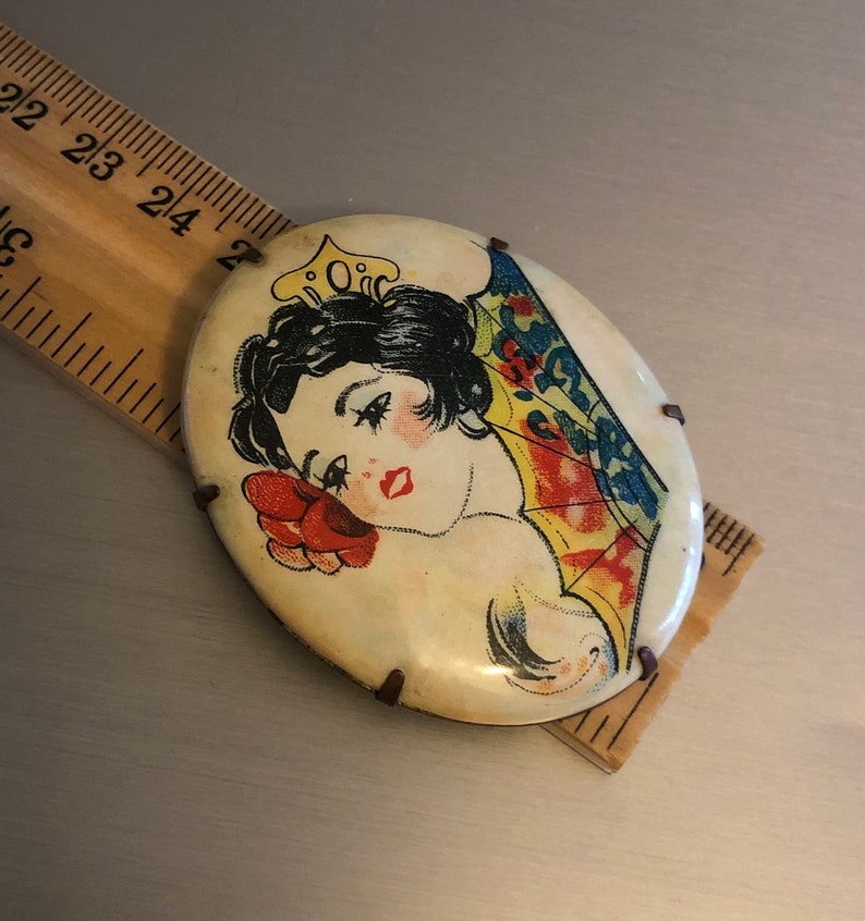 Pu&ograve; includere: Una spilla vintage di forma ovale con un'immagine colorata di una donna con una corona e un ventaglio. La spilla &egrave; in metallo e ha uno sfondo bianco.