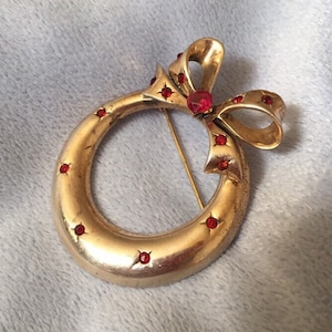 Könnte beinhalten: Eine goldfarbene Brosche in Form eines Kranzes mit einer roten Schleife. Der Kranz ist mit roten Strasssteinen verziert.