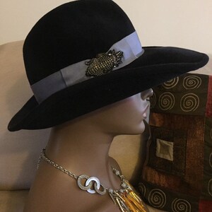 May include: A black felt fedora hat with a wide brim and a gray ribbon. The hat has a decorative pin with black and silver accents.