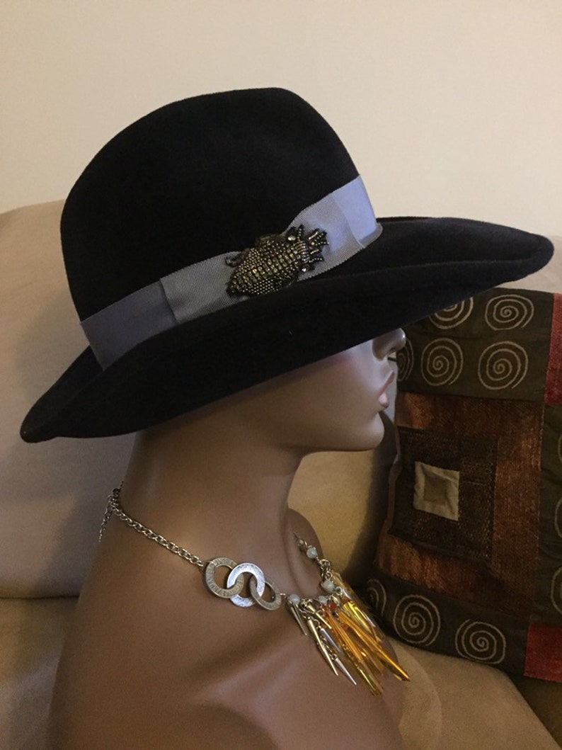 May include: A black felt fedora hat with a wide brim and a gray ribbon. The hat has a decorative pin with black and silver accents.