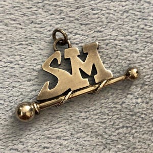 Puede incluir: Colgante plateado con las letras "SM" y una barra decorativa con extremos de bola. El colgante tiene un lazo para sujetarlo a una cadena o pulsera. El diseño es sencillo y elegante, adecuado para joyería.