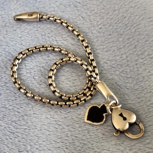 May include: Silver necklace with a chain-link design. The necklace has a heart-shaped padlock charm and a black heart-shaped charm. The necklace is displayed on a light blue surface.