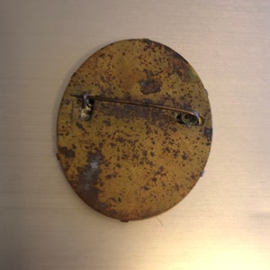 Pu&ograve; includere: Il retro di uno spillo rotondo, di forma ovale, in ottone con un fermaglio a spillo. Lo spillo &egrave; ossidato e ha un aspetto usurato e invecchiato.