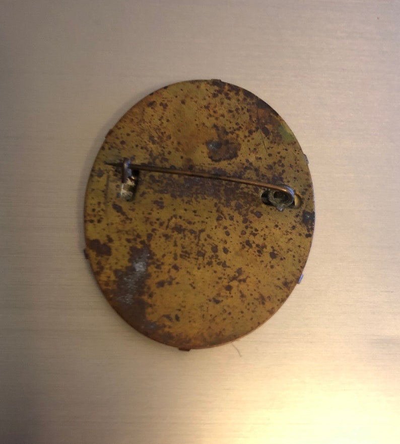 Pu&ograve; includere: Il retro di uno spillo rotondo, di forma ovale, in ottone con un fermaglio a spillo. Lo spillo &egrave; ossidato e ha un aspetto usurato e invecchiato.