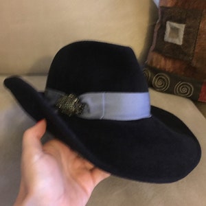 May include: A black felt fedora hat with a wide brim and a light blue ribbon. The hat has a decorative embellishment on the front.