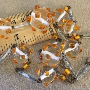 Könnte beinhalten: Eine Vintage-Halskette mit durchsichtigen Glasperlen mit weißen Streifen und orangefarbenen Punkten. Die Perlen sind durch eine silberne Kette verbunden.
