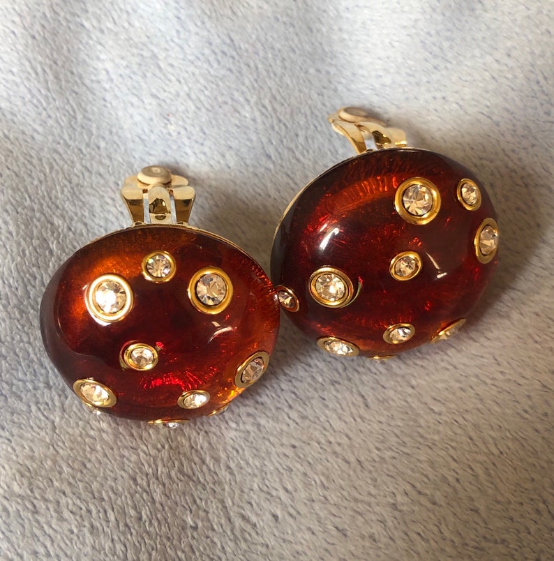 May include: A pair of gold tone clip-on earrings with a brown, round, dome-shaped design. Each earring is decorated with clear rhinestones.