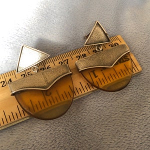 Puede incluir: Un par de pendientes geométricos de ámbar y plata. Los pendientes tienen forma de semicírculo con un triángulo de plata en la parte superior. Los pendientes se miden con una regla.
