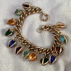 Op de afbeelding: Een gouden kettingarmband met meerdere hartvormige bedels. Elke bedel heeft een anders gekleurde glazen steen erin. De stenen zijn rood, oranje, geel, groen, blauw en paars.