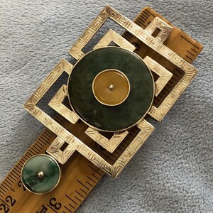 Puede incluir: Broche plateado con diseño geométrico, con una piedra circular grande de color verde oscuro con una piedra central más pequeña de color marrón claro. Una piedra verde más pequeña a juego está en el lateral. El broche está sobre una regla de madera.