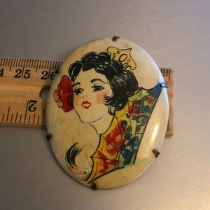 Pu&ograve; includere: Broche vintage di forma ovale con un'immagine colorata di una donna con i capelli scuri che indossa un vestito a motivi gialli e blu e un fiore rosso dietro l'orecchio. La broche ha uno sfondo bianco ed &egrave; decorata con accenti metallici.