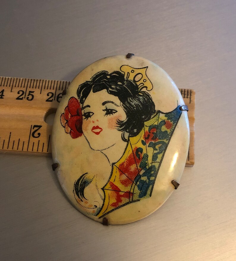 Pu&ograve; includere: Broche vintage di forma ovale con un'immagine colorata di una donna con i capelli scuri che indossa un vestito a motivi gialli e blu e un fiore rosso dietro l'orecchio. La broche ha uno sfondo bianco ed &egrave; decorata con accenti metallici.