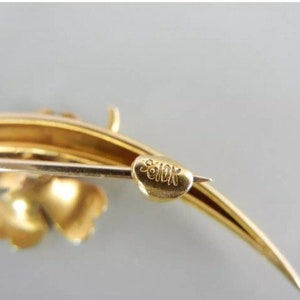 May include: A gold-toned brooch with a clover design and a curved stem. The back of the brooch is marked "9010K".
