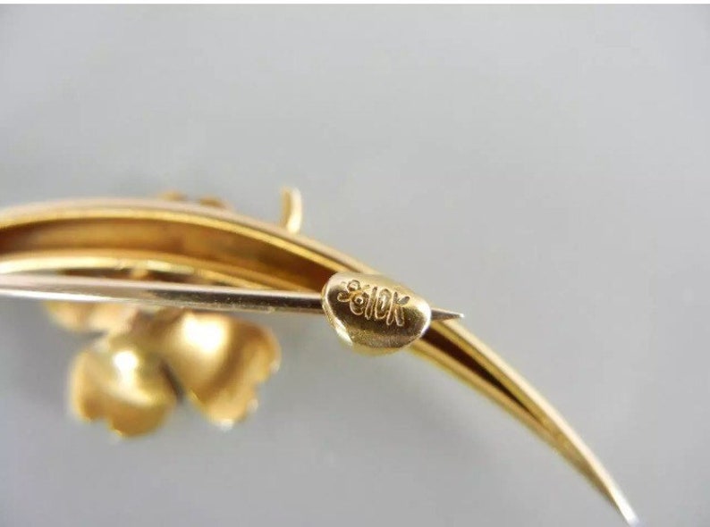 May include: A gold-toned brooch with a clover design and a curved stem. The back of the brooch is marked "9010K".