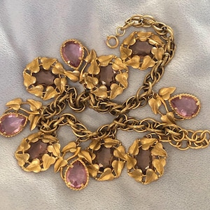 Op de afbeelding: Een gouden ketting met een bladontwerp en paarse glazen stenen. De ketting is gemaakt van metaal en heeft een delicate, vrouwelijke uitstraling.