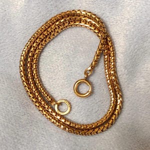 May include: A gold-colored necklace with a snake chain design. The necklace is made of small, interlocking links that create a smooth, flexible chain. The necklace has a circular clasp and a small ring at the end.